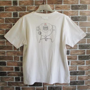 May include: White t-shirt with a black and white graphic of a video game controller on the back. The t-shirt is hanging on a white hanger against a brick wall.