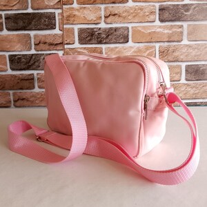 Puede incluir: Bolso bandolera rosa claro de forma rectangular. El bolso tiene cierre de cremallera y una correa ajustable rosa a juego. El bolso est&aacute; hecho de un material liso. Fondo de pared de ladrillo.