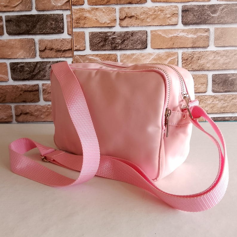 Puede incluir: Bolso bandolera rosa claro de forma rectangular. El bolso tiene cierre de cremallera y una correa ajustable rosa a juego. El bolso est&aacute; hecho de un material liso. Fondo de pared de ladrillo.