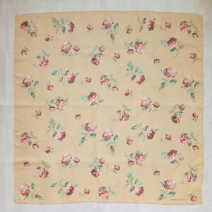 Op de afbeelding: Een vierkante sjaal in een zandkleur, versierd met een herhalend bloemenpatroon. De bloemen zijn een mix van rood en roze, met groene bladeren. De sjaal is gemaakt van een zachte stof en heeft de tekst "LAURA ASHLEY" in de rechterbenedenhoek.