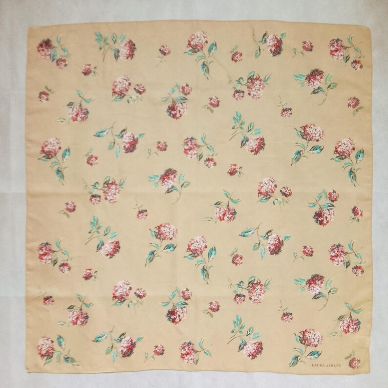 Op de afbeelding: Een vierkante sjaal in een zandkleur, versierd met een herhalend bloemenpatroon. De bloemen zijn een mix van rood en roze, met groene bladeren. De sjaal is gemaakt van een zachte stof en heeft de tekst "LAURA ASHLEY" in de rechterbenedenhoek.
