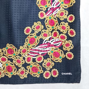 May include: A navy blue silk scarf with a repeating pattern. The scarf features a design of gold-colored floral shapes with pink and clear gemstones. The word "CHANEL" is printed in white in the lower right corner.