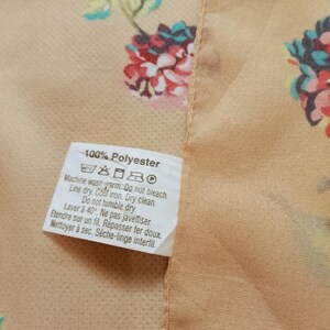 Op de afbeelding: Close-up van een perzik-kleurige stof met een bloemenpatroon in roze, rood en groen. Een wit label toont wasvoorschriften en de tekst "100% Polyester". De stof heeft een gladde textuur.