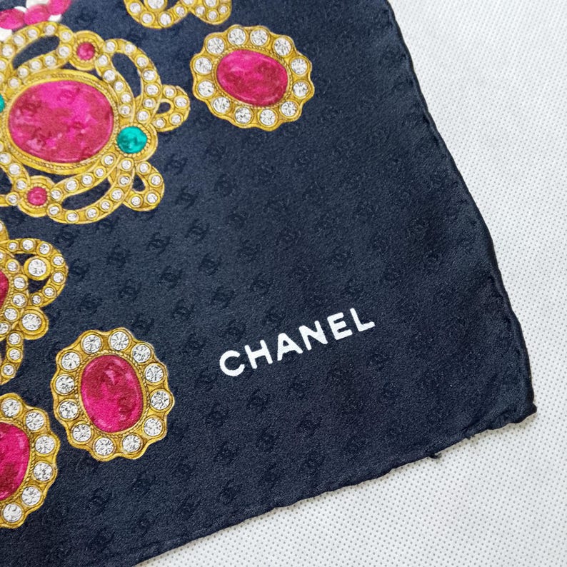 May include: A black silk scarf with a repeating pattern of interlocking Cs and jewel-like embellishments. The scarf features gold-colored frames with pink and clear gemstones. The word "CHANEL" is printed in white.