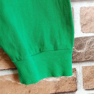 May include: Close-up of a bright green garment sleeve with a ribbed cuff. The fabric has a textured, honeycomb-like pattern. The sleeve is against a brick wall.