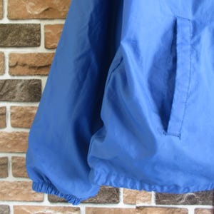 Pode incluir: Uma jaqueta corta-vento azul com fecho de correr e um bolso do lado direito. A jaqueta tem um cord&atilde;o na parte inferior.