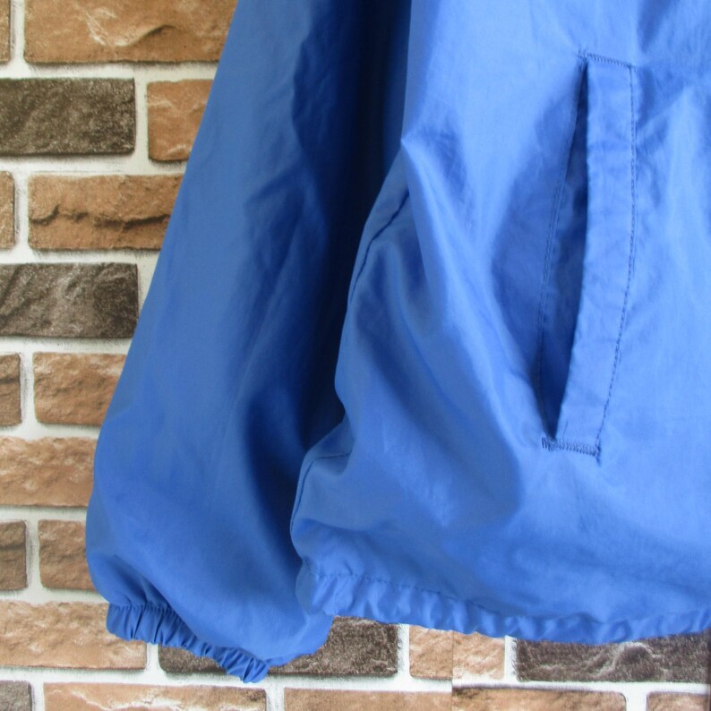 Pode incluir: Uma jaqueta corta-vento azul com fecho de correr e um bolso do lado direito. A jaqueta tem um cord&atilde;o na parte inferior.