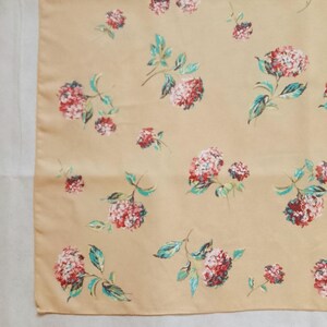 Op de afbeelding: Een beige stof met een bloemenprint. Het ontwerp toont roze en rode bloemclusters met groene bladeren. De bloemen zijn verspreid over de stof, wat een delicate en vrouwelijke uitstraling cre&euml;ert. De stof lijkt glad, mogelijk zijde of satijn.