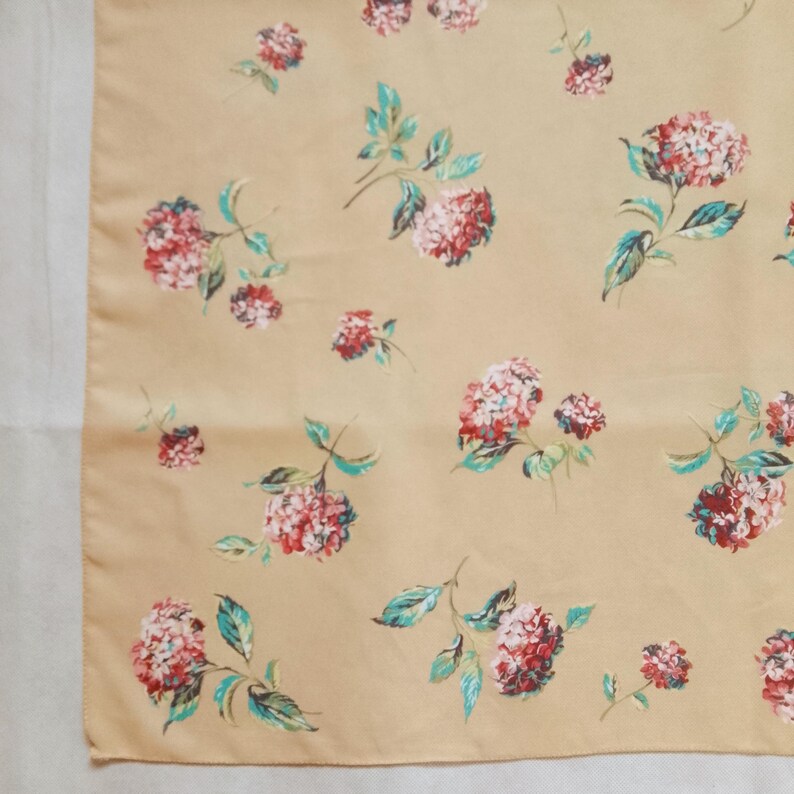 Op de afbeelding: Een beige stof met een bloemenprint. Het ontwerp toont roze en rode bloemclusters met groene bladeren. De bloemen zijn verspreid over de stof, wat een delicate en vrouwelijke uitstraling cre&euml;ert. De stof lijkt glad, mogelijk zijde of satijn.