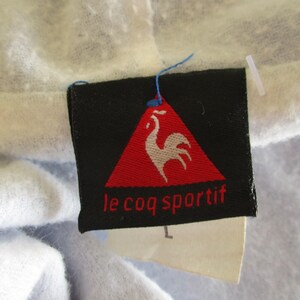 Pode incluir: Um r&oacute;tulo preto e vermelho com um logotipo de galo branco e o texto "le coq sportif".