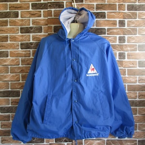 Pode incluir: Uma jaqueta azul com capuz, forro branco e um logotipo de galo vermelho. O logotipo est&aacute; em um tri&acirc;ngulo branco com o texto "le coq sportif" abaixo.