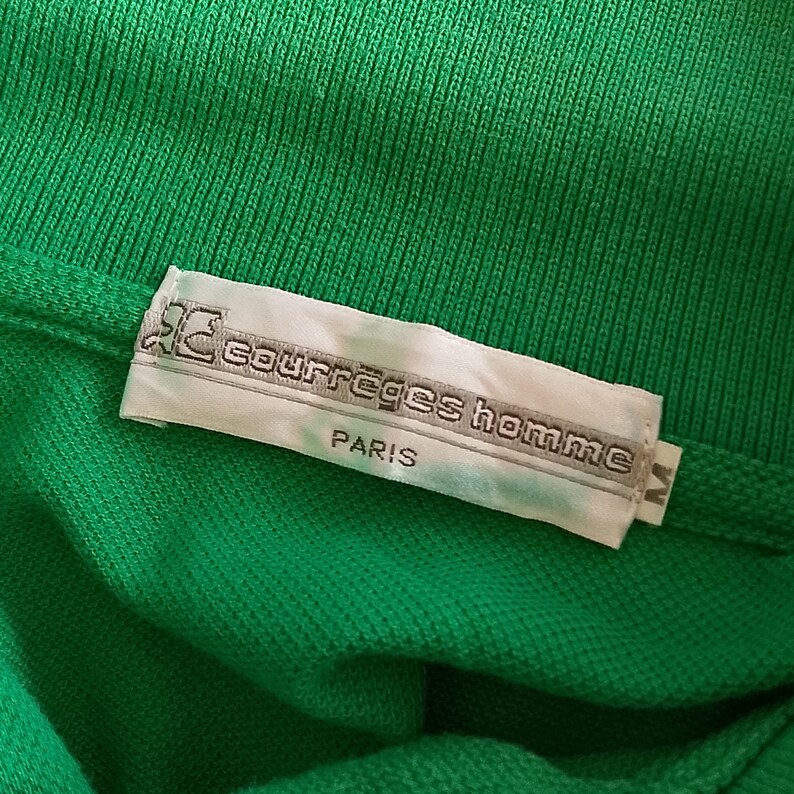 May include: Close-up of a bright green polo shirt with a white label. The label reads "courr&egrave;ges homme PARIS" in a gray font. The shirt is made of a textured fabric.