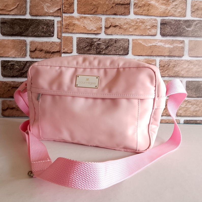 Puede incluir: Un bolso bandolera Courr&egrave;ges rosa claro con una correa ajustable a juego. El bolso tiene forma rectangular con un bolsillo frontal y una peque&ntilde;a placa dorada con el nombre de la marca. El bolso est&aacute; hecho de un material liso, posiblemente nailon.