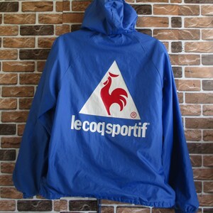 Pode incluir: Uma jaqueta azul com capuz com um logotipo triangular branco que apresenta um galo vermelho e o texto "le coq sportif" em branco.
