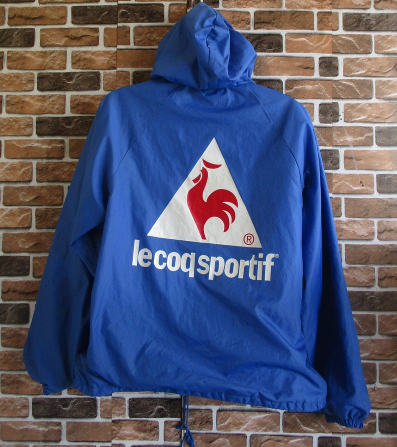 Pode incluir: Uma jaqueta azul com capuz com um logotipo triangular branco que apresenta um galo vermelho e o texto "le coq sportif" em branco.