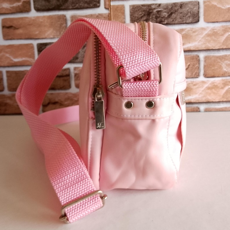 Puede incluir: Bolso bandolera rosa claro con una correa ajustable a juego. El bolso tiene un compartimento principal con cremallera y un bolsillo delantero m&aacute;s peque&ntilde;o. Los herrajes son plateados y el bolso est&aacute; hecho de un material liso, posiblemente nailon.