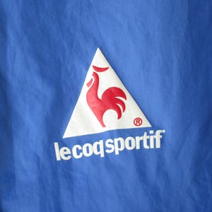 Pode incluir: Uma jaqueta azul com um logotipo triangular branco que apresenta um galo vermelho e o texto "le coq sportif" em branco.