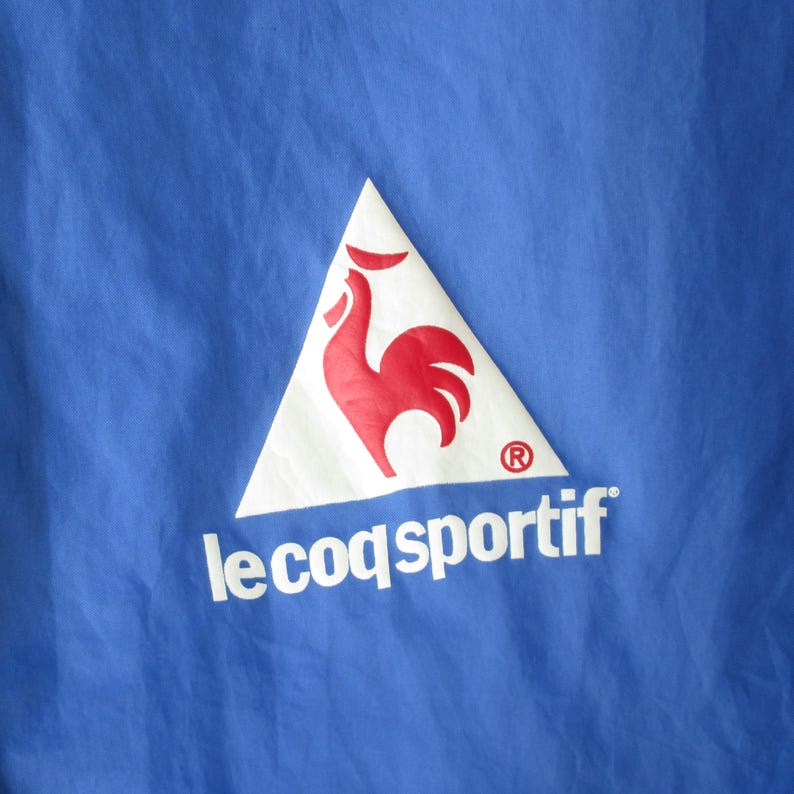 Pode incluir: Uma jaqueta azul com um logotipo triangular branco que apresenta um galo vermelho e o texto "le coq sportif" em branco.
