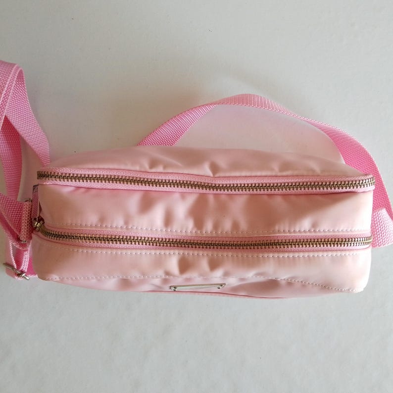 Puede incluir: Un bolso de cintura rosa claro con dos compartimentos con cremallera y una correa ajustable a juego. La bolsa tiene una peque&ntilde;a etiqueta met&aacute;lica de color plateado en la parte delantera. Las cremalleras son de color bronce.