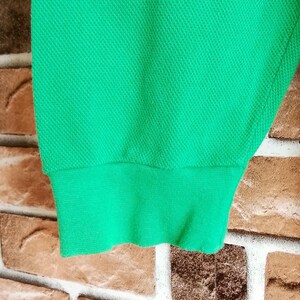 May include: Close-up of a bright green fabric sleeve with a ribbed cuff. The fabric has a textured, woven appearance. The sleeve is against a brick wall background.