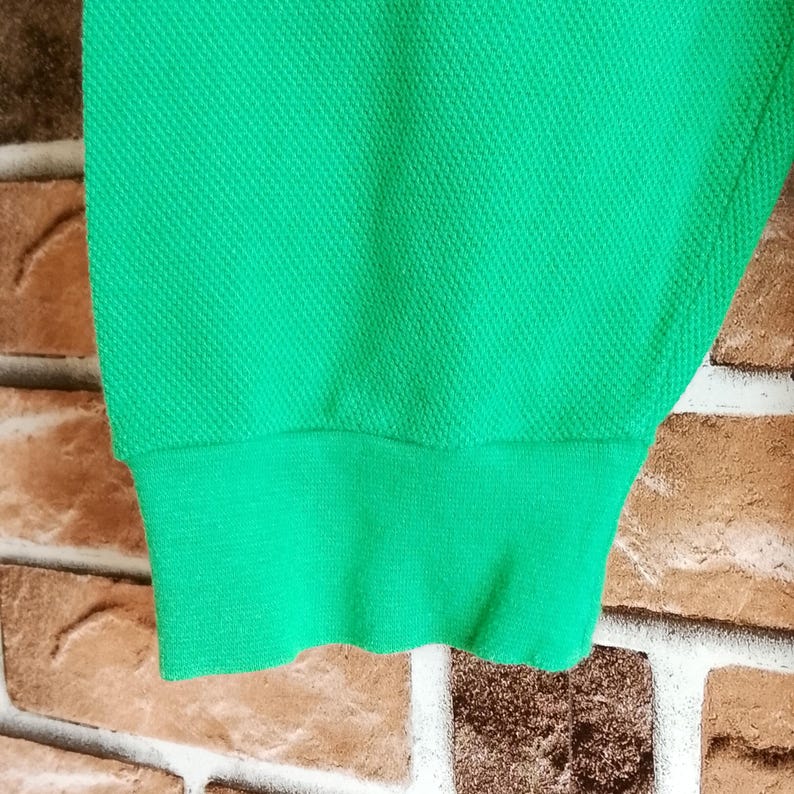 May include: Close-up of a bright green fabric sleeve with a ribbed cuff. The fabric has a textured, woven appearance. The sleeve is against a brick wall background.