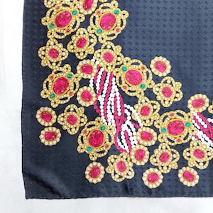 May include: A black silk scarf with an ornate design. Gold-colored filigree frames large, oval, pink gemstones, accented with small white and green stones. White and pink beaded strands add detail to the corner.