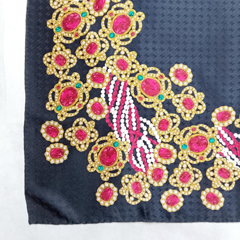 May include: A black silk scarf with an ornate design. Gold-colored filigree frames large, oval, pink gemstones, accented with small white and green stones. White and pink beaded strands add detail to the corner.