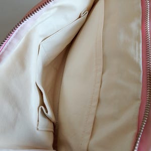 Puede incluir: Interior de un bolso rosa con forro crema y m&uacute;ltiples bolsillos. La cremallera es plateada. El interior del bolso es visible, mostrando los compartimentos y los detalles de las costuras.