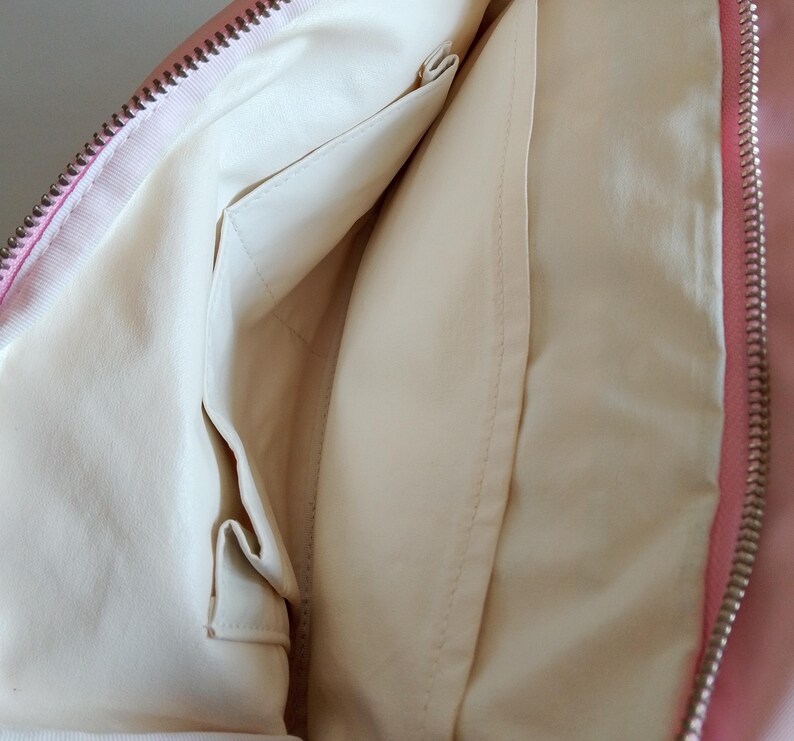 Puede incluir: Interior de un bolso rosa con forro crema y m&uacute;ltiples bolsillos. La cremallera es plateada. El interior del bolso es visible, mostrando los compartimentos y los detalles de las costuras.