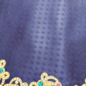 May include: A navy blue textile with a repeating pattern of interlocking Cs. The lower left corner features a gold-colored, ornate embellishment with green, pink, and clear gemstones. The textile has a subtle sheen.