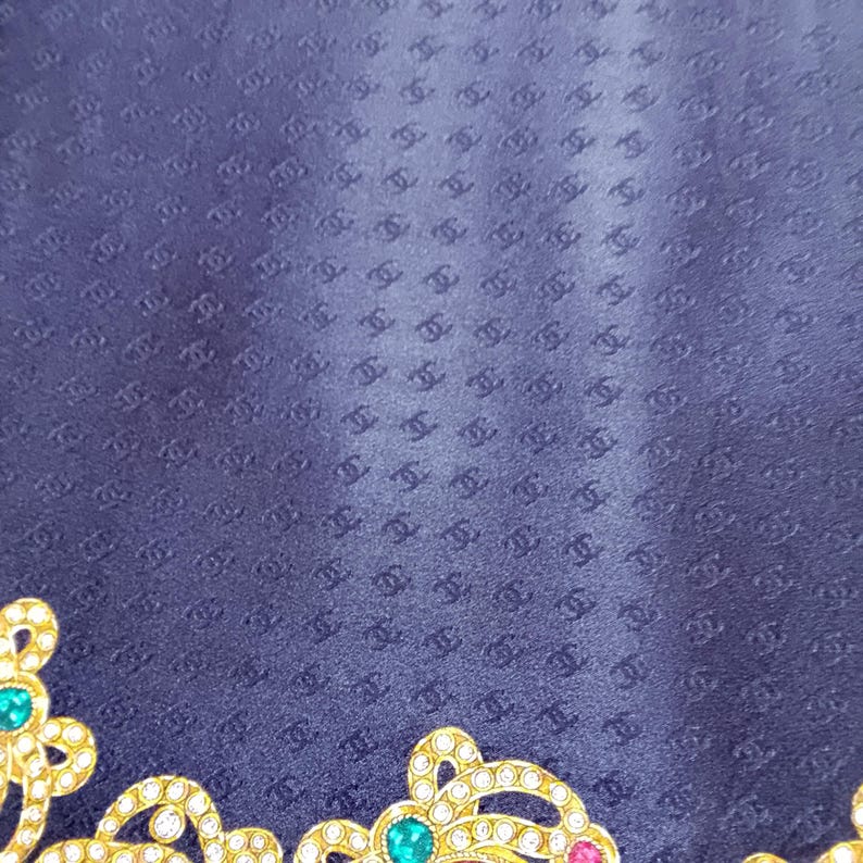 May include: A navy blue textile with a repeating pattern of interlocking Cs. The lower left corner features a gold-colored, ornate embellishment with green, pink, and clear gemstones. The textile has a subtle sheen.