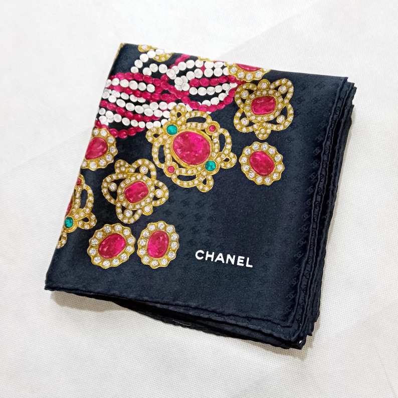 May include: Black silk scarf with a repeating pattern of the Chanel logo. The scarf features a design of gold-colored floral shapes with pink and clear gemstones, and white pearl-like beads. The word "CHANEL" is printed in white.