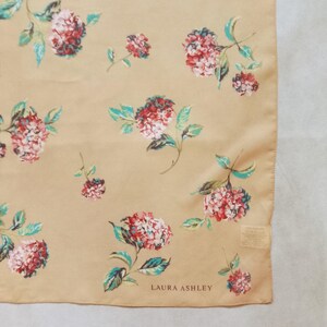 Op de afbeelding: Een vierkante sjaal in beige met een bloemenpatroon. Het ontwerp toont clusters van rode en witte bloemen met turquoise bladeren. De merknaam "LAURA ASHLEY" staat onderaan gedrukt.