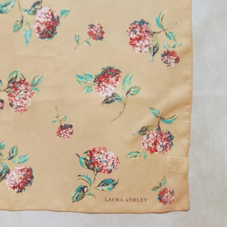 Op de afbeelding: Een vierkante sjaal in beige met een bloemenpatroon. Het ontwerp toont clusters van rode en witte bloemen met turquoise bladeren. De merknaam "LAURA ASHLEY" staat onderaan gedrukt.