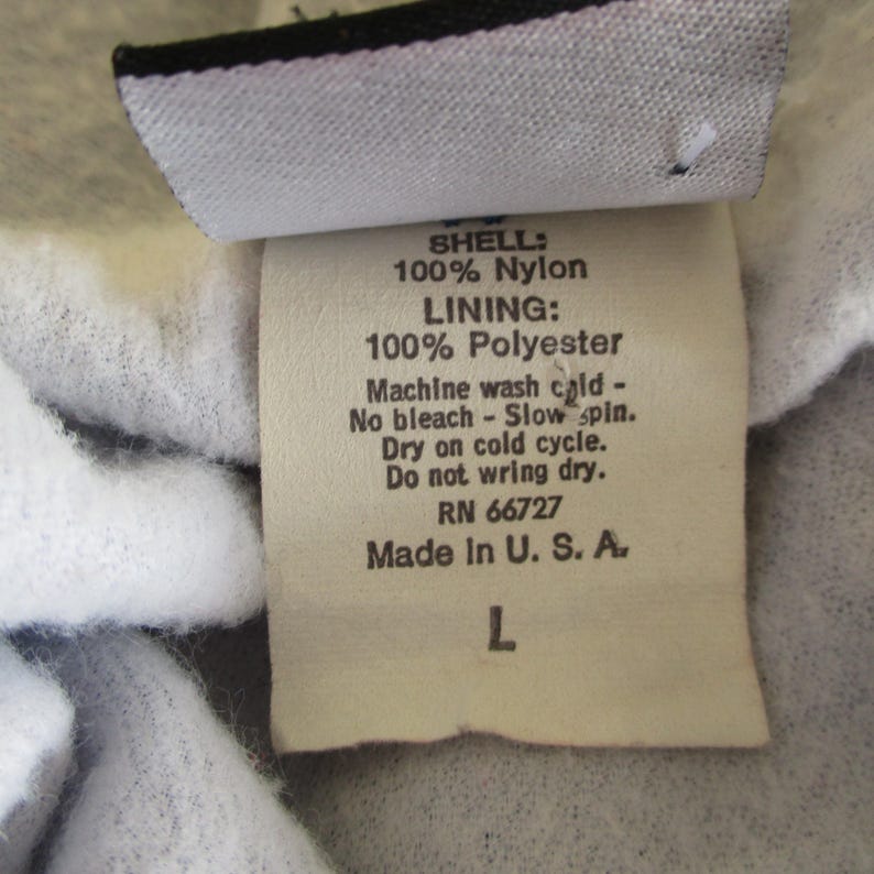 Pode incluir: Uma etiqueta branca com texto preto que detalha a composi&ccedil;&atilde;o do tecido e as instru&ccedil;&otilde;es de cuidado para uma pe&ccedil;a de roupa. A etiqueta diz "SHELL: 100% Nylon LINING: 100% Polyester Machine wash cold - No bleach - Slow spin. Dry on cold cycle. Do not wring dry. RN 66727 Made In U. S. A. L".