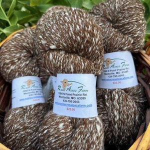May include: Three skeins of brown and white alpaca yarn in a wicker basket. The yarn is speckled with white flecks and has a rustic, natural look. The yarn is labeled "Rush Acres Farm" with contact information and a price of $22.00.