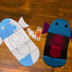 May include: Two paper fish cutouts, one blue and white, the other blue, black, and red. The blue and white fish is partially colored. Two small paper figures are next to the fish. The background is a wooden surface.