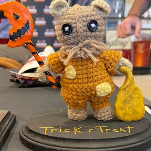 Puede incluir: Un oso de crochet marrón que lleva un disfraz amarillo y marrón con lunares naranja y negro. El oso sostiene un palo con una calabaza tallada en la parte superior. El oso está de pie sobre una base negra con las palabras "Trick or Treat" escritas en amarillo.