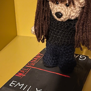 Puede incluir: Un oso de crochet con pelo largo marrón de pie sobre un libro negro titulado "MALIGNANT" de "EMILY MAY".