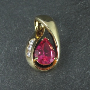 May include: A gold pendant with a teardrop-shaped, bright red gemstone. The pendant has a curved design with three small, clear stones on the side. The jewelry is set against a dark gray background.