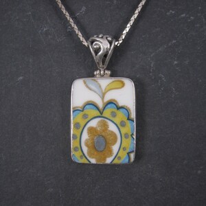 May include: A silver chain necklace with a rectangular pendant featuring a floral design in shades of yellow, blue, and gray on a white background.
