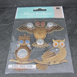 May include: A package of three dimensional owl stickers. The owls are metallic gold with jewel-toned eyes and silver accents. One owl has outstretched wings, another has a crown, and the third is perched on a branch. The package reads "Jolee's Boutique".