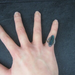 May include: A silver ring with an oval, iridescent blue-green stone. The ring has a decorative, twisted rope design around the stone and on the sides. The ring is on a hand, with a dark background.