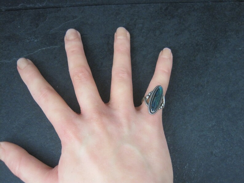 May include: A silver ring with an oval, iridescent blue-green stone. The ring has a decorative, twisted rope design around the stone and on the sides. The ring is on a hand, with a dark background.
