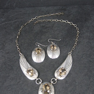 May include: A silver necklace and earrings set with a brown gemstone. The necklace features a pendant with a large oval-shaped gemstone and two smaller gemstones on each side. The earrings are simple and feature a single oval-shaped gemstone.