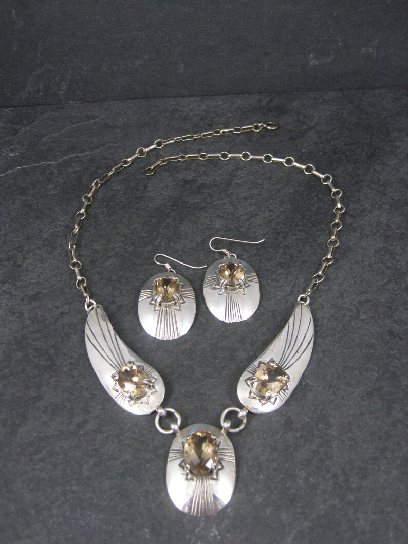 May include: A silver necklace and earrings set with a brown gemstone. The necklace features a pendant with a large oval-shaped gemstone and two smaller gemstones on each side. The earrings are simple and feature a single oval-shaped gemstone.