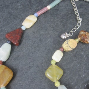 May include: A multi-colored gemstone necklace with a silver clasp. The necklace features various shapes and sizes of stones, including red, green, yellow, and brown.