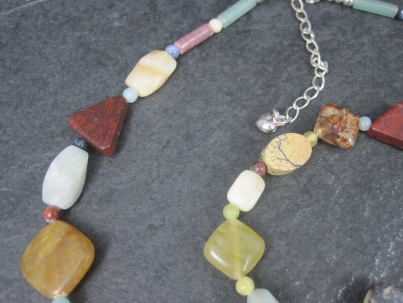 May include: A multi-colored gemstone necklace with a silver clasp. The necklace features various shapes and sizes of stones, including red, green, yellow, and brown.