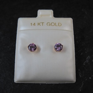 May include: A pair of stud earrings with round, light purple gemstones set in gold. The earrings are displayed on a white card that reads "14 KT. GOLD". The background is a dark, textured surface.