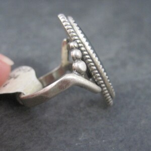 May include: A silver ring with an oval, dark-colored stone. The ring has a decorative, textured border and small, round accents. The ring is shown from a side angle, highlighting its design and craftsmanship. The ring is a piece of jewelry.